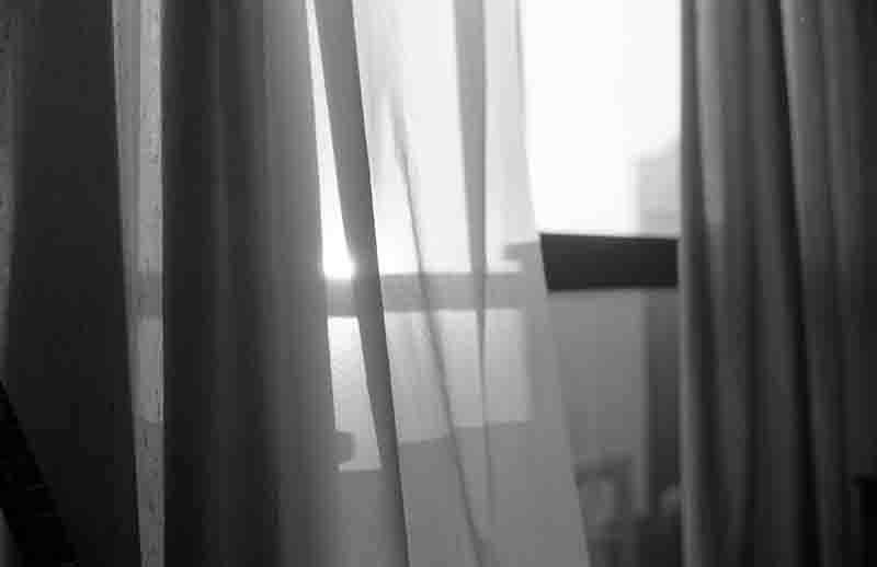 Ein Schwarz-Weiß-Foto, das durch transparente, weiche Vorhänge hindurch den Blick auf ein helles Fenster freigibt, wobei das Sonnenlicht von hinten durchscheint und feine Kontraste erzeugt.